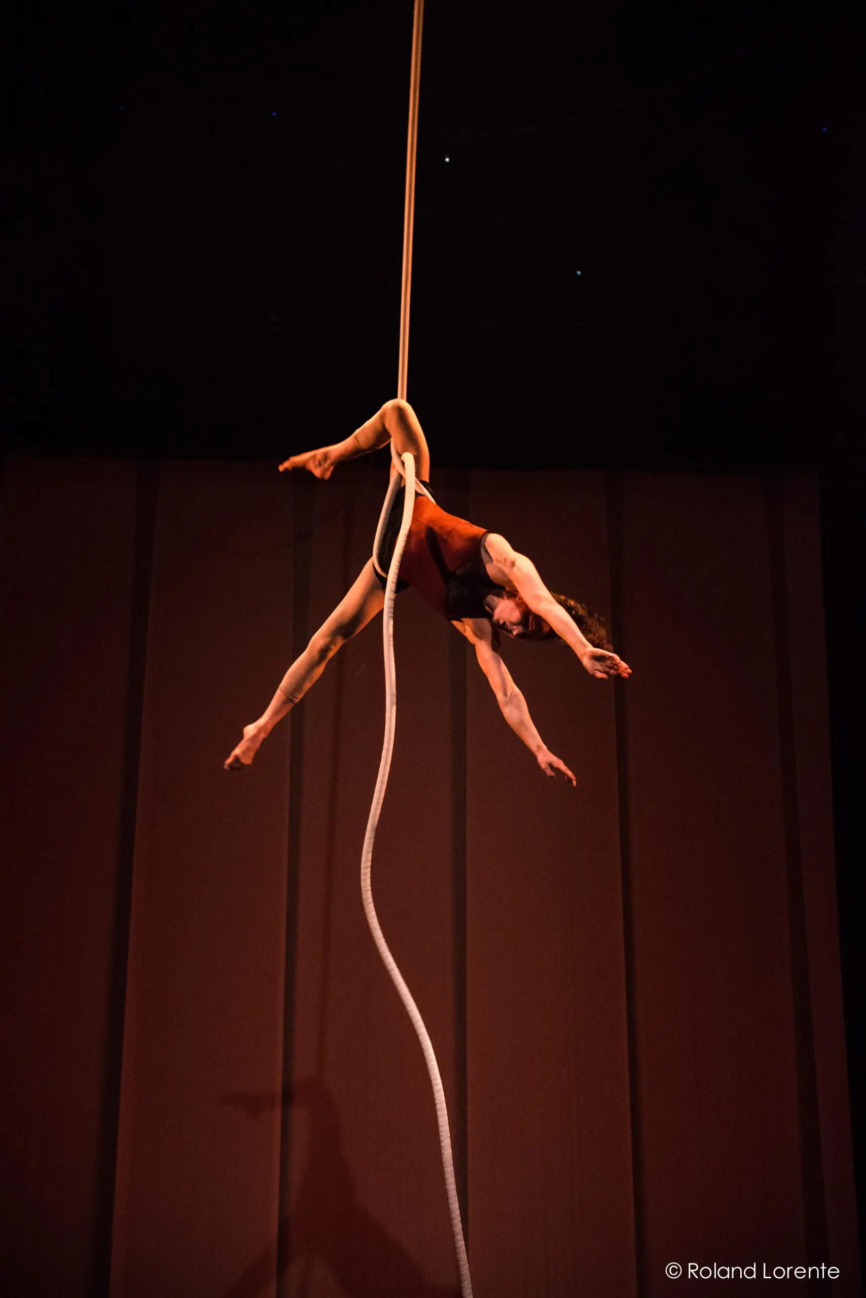Rosita Hendry, une acrobate aérienne, se produit au-dessus de la scène, suspendue à l'envers à une corde blanche, les bras et les jambes étendus gracieusement dans une pose dramatique sur une toile de fond sombre.