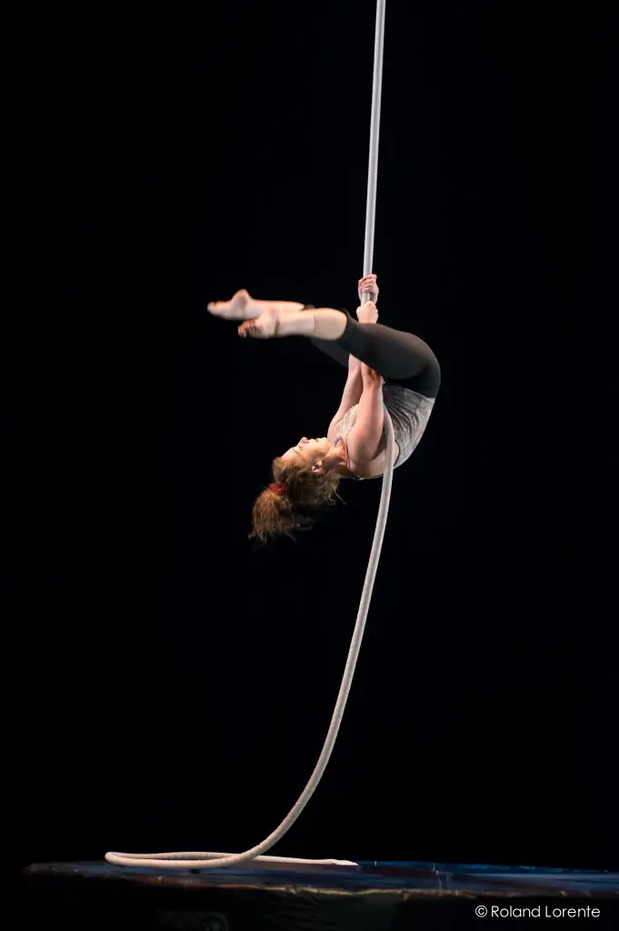 Une artiste aérienne, Una Bennett, est suspendue la tête en bas à une corde blanche, arquant gracieusement son dos et étendant ses jambes sur un fond sombre.