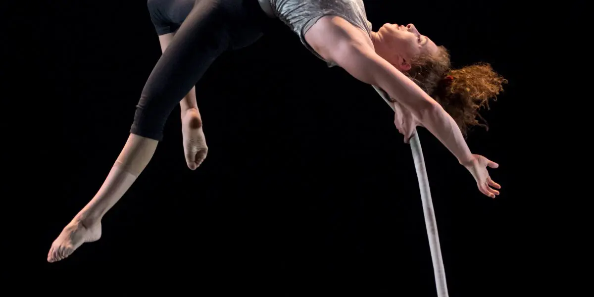 Una Bennett exécute une routine aérienne, en arquant le dos et en se suspendant à l'envers à une corde blanche sur un fond noir, les bras tendus et les jambes pliées.