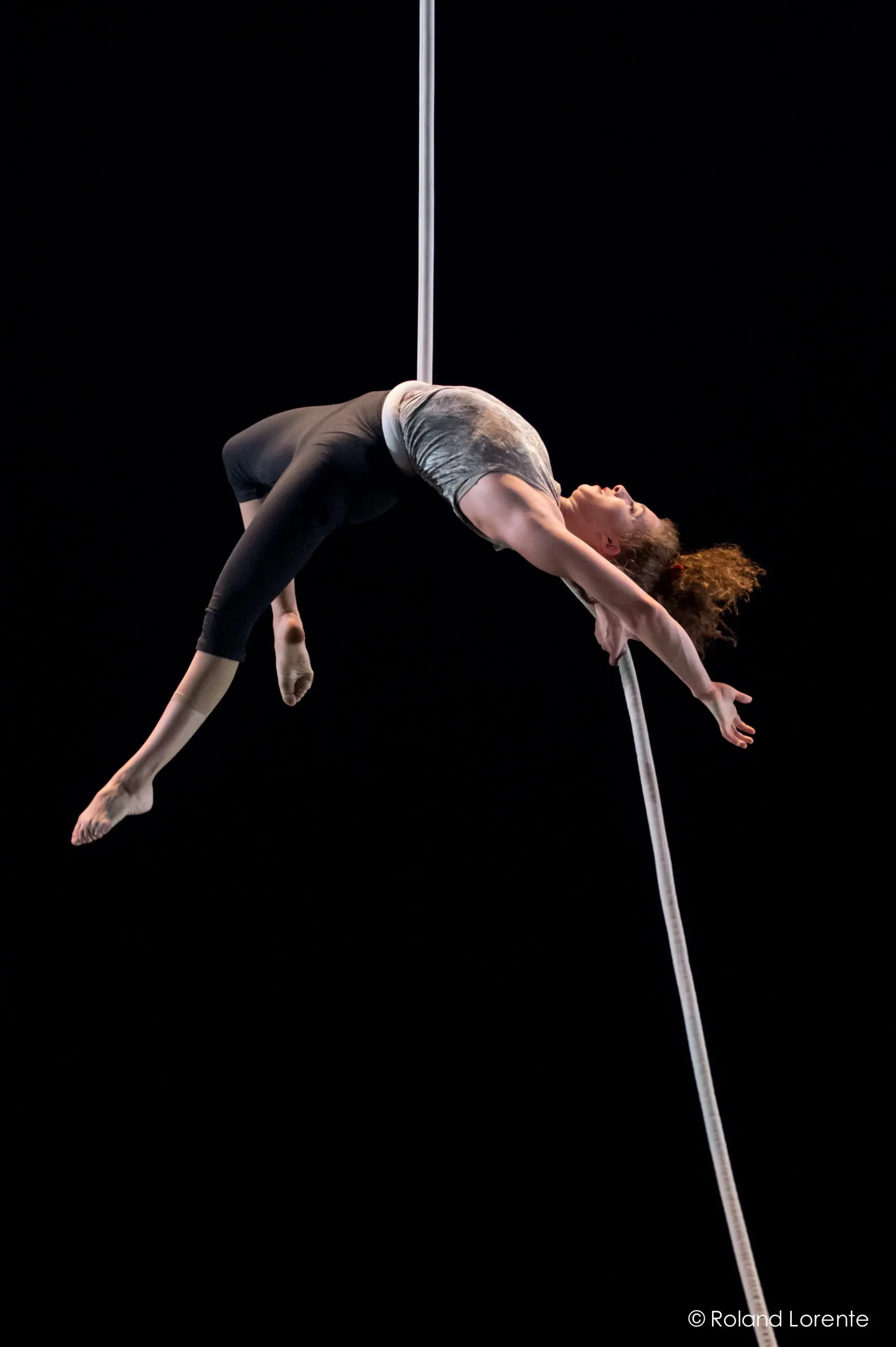 Una Bennett exécute une routine aérienne, en arquant le dos et en se suspendant à l'envers à une corde blanche sur un fond noir, les bras tendus et les jambes pliées.