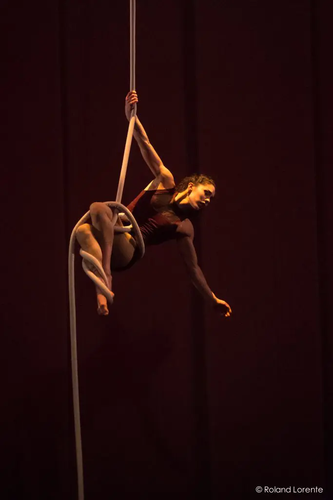 Une artiste aérienne, Rosita Hendry, vêtue d'une tenue marron, pose gracieusement dans les airs, enveloppée dans des soies aériennes blanches sur un fond sombre. L'éclairage chaud de la scène met en valeur son expression concentrée et sa forme athlétique.