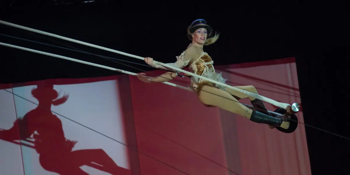 Un artiste de cirque en costume et chapeau dorés se balance sur un trapèze, projetant une ombre sur un fond rouge et blanc, lors de la performance scénique envoûtante de Roxane Gilliand.