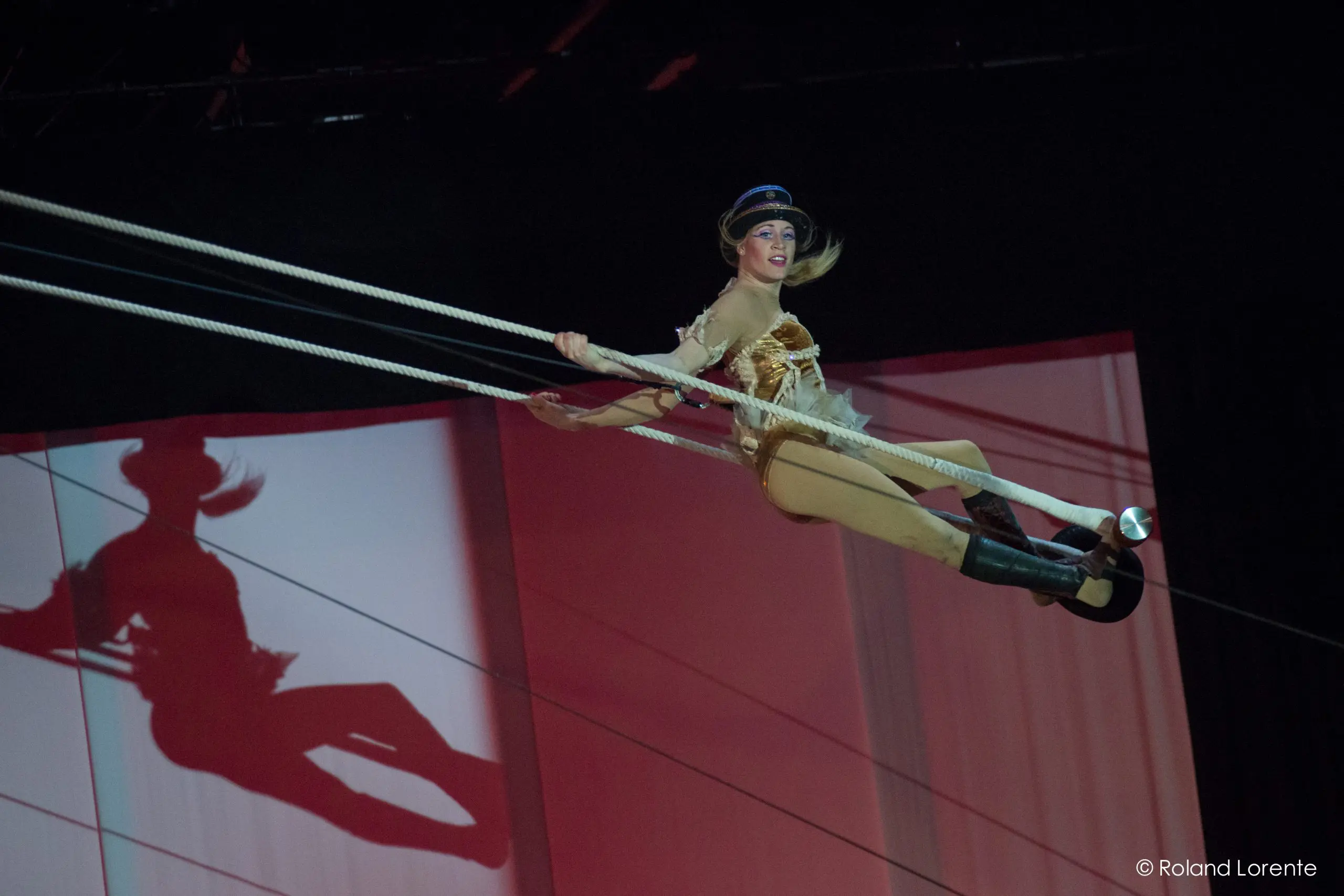 Un artiste de cirque en costume et chapeau dorés se balance sur un trapèze, projetant une ombre sur un fond rouge et blanc, lors de la performance scénique envoûtante de Roxane Gilliand.