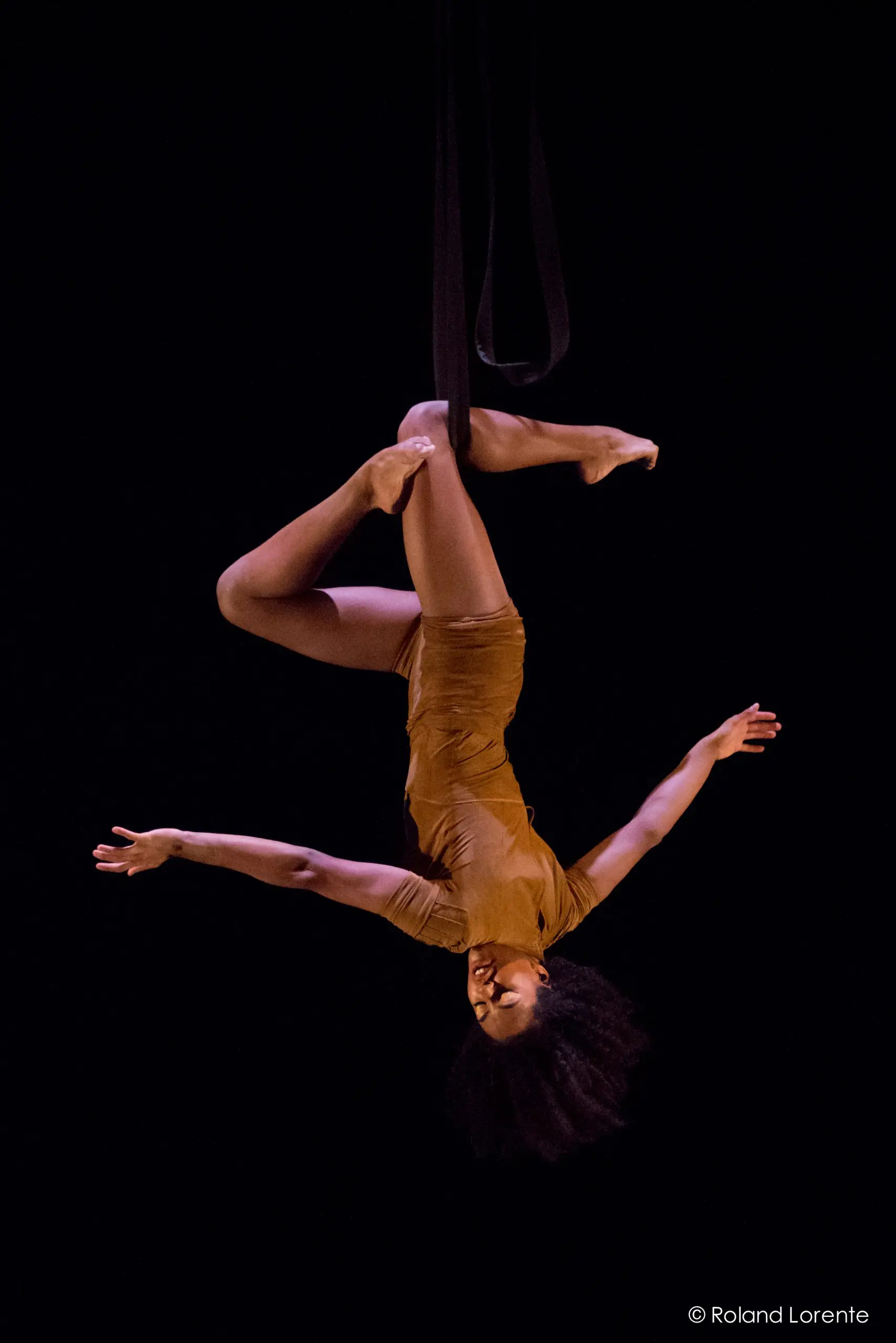 L'artiste aérienne Tuedon Ariri, vêtue d'une tenue marron, est suspendue par les jambes à une sangle aérienne noire, les bras gracieusement étendus, sur un fond sombre.