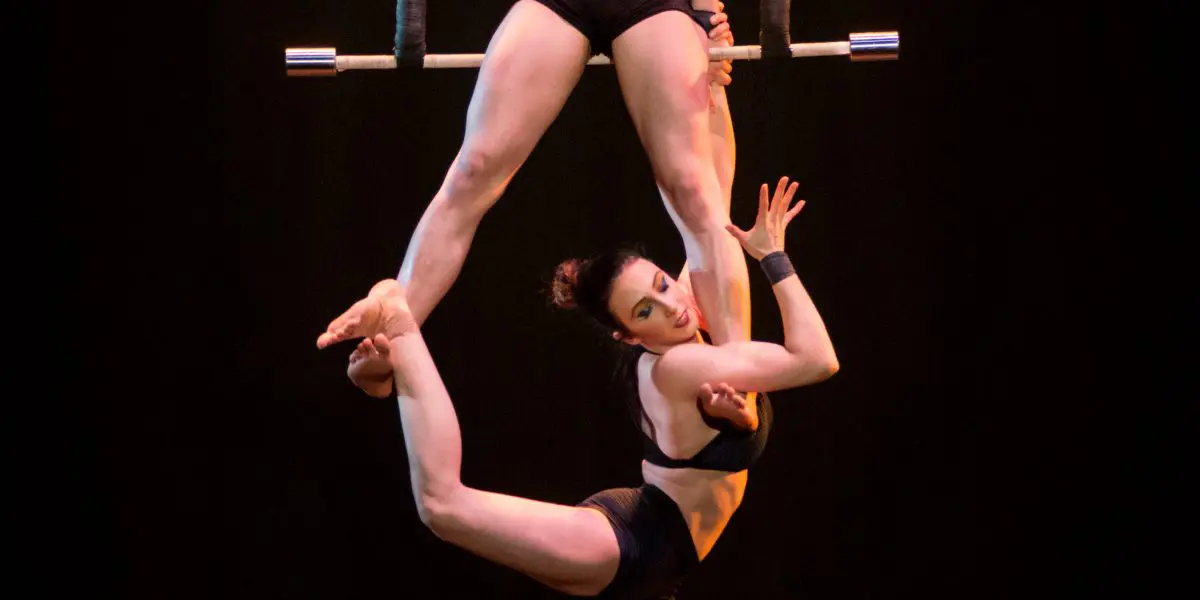 Deux acrobates féminines en tenue noire exécutent un numéro aérien sur un trapèze, l'une assise et tenant la barre tandis que Virginie Gerbeau est suspendue à l'envers, agrippée à la jambe de sa partenaire. L'arrière-plan est sombre et l'éclairage dramatique.