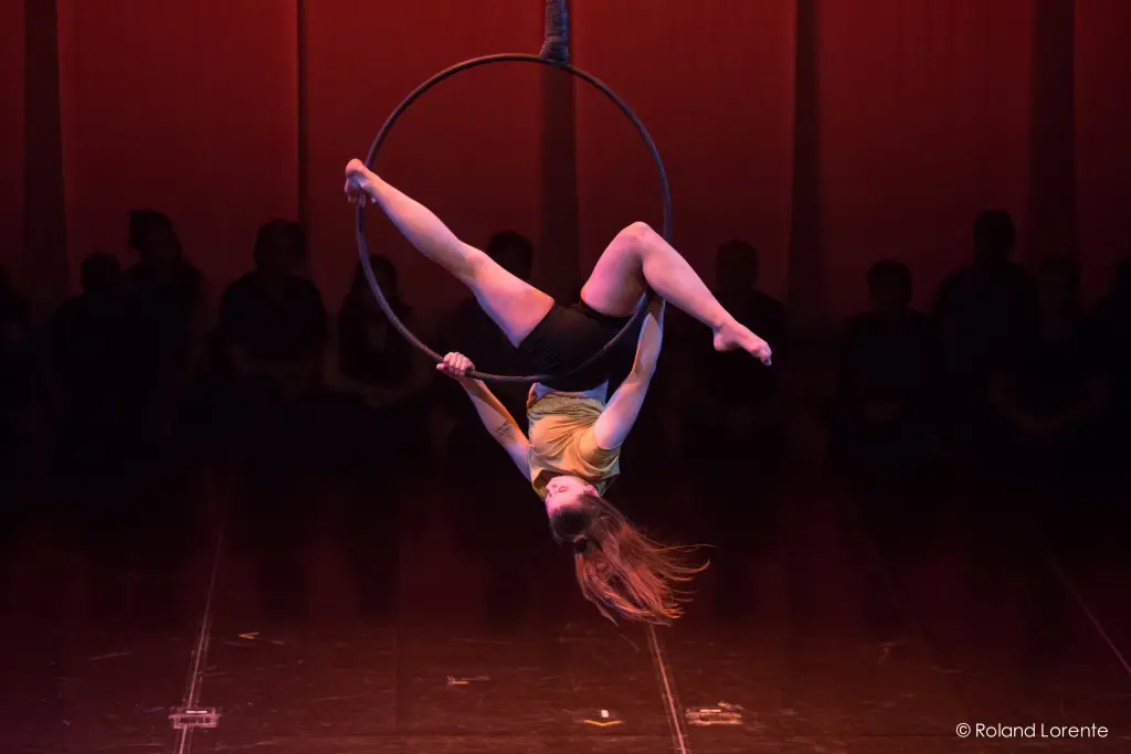 Éliza Gélinas-Lance, une artiste aérienne, est suspendue à l'envers à un cerceau, ses jambes s'agrippant au sommet et ses cheveux tombant vers le sol, sur une scène faiblement éclairée à l'arrière-plan rougeâtre.