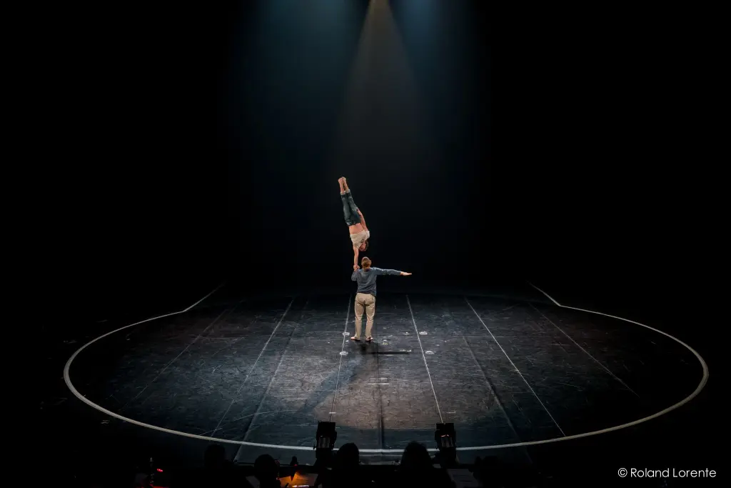 Deux acrobates, Julius Bitterling et César Mispelon, évoluent sur une scène sombre ; l'un soulève l'autre au-dessus de sa tête à l'aide de ses deux mains. Un projecteur les éclaire, mettant en valeur leur équilibre et leur habileté à l'intérieur d'une aire de spectacle circulaire.