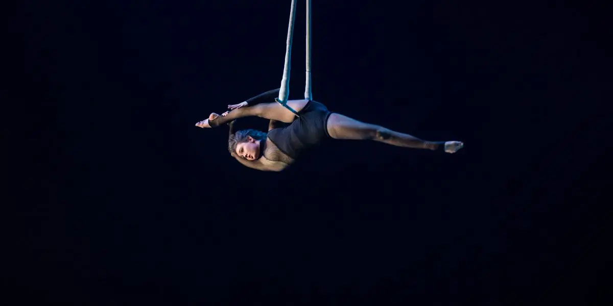 Une voltigeuse, Georgia Simpson, vêtue d'un justaucorps noir, exécute une pose acrobatique tout en étant suspendue horizontalement à des sangles aériennes sur un fond de scène sombre.
