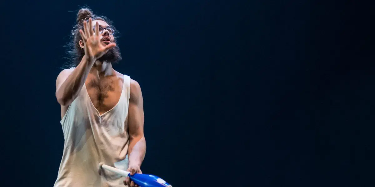 Un artiste pieds nus, rappelant Kellin-Quinn Hentoff-Killian, vêtu d'une tenue blanche ample, jongle avec des massues bleues sur une scène sombre. Un survêtement est posé à proximité, tandis qu'un éclairage dramatique met en valeur la concentration intense et les mouvements fluides du jongleur.