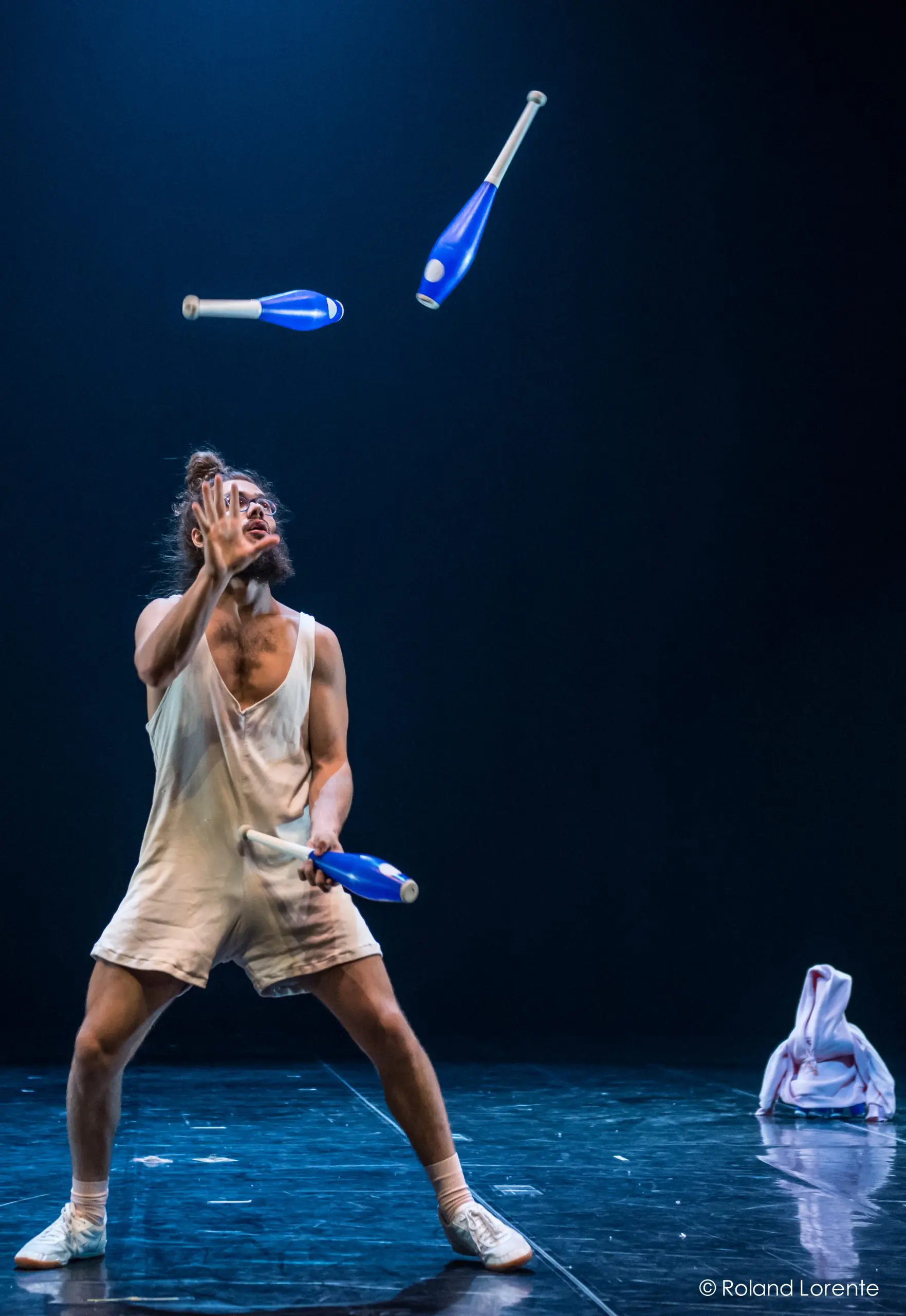 Un artiste pieds nus, rappelant Kellin-Quinn Hentoff-Killian, vêtu d'une tenue blanche ample, jongle avec des massues bleues sur une scène sombre. Un survêtement est posé à proximité, tandis qu'un éclairage dramatique met en valeur la concentration intense et les mouvements fluides du jongleur.