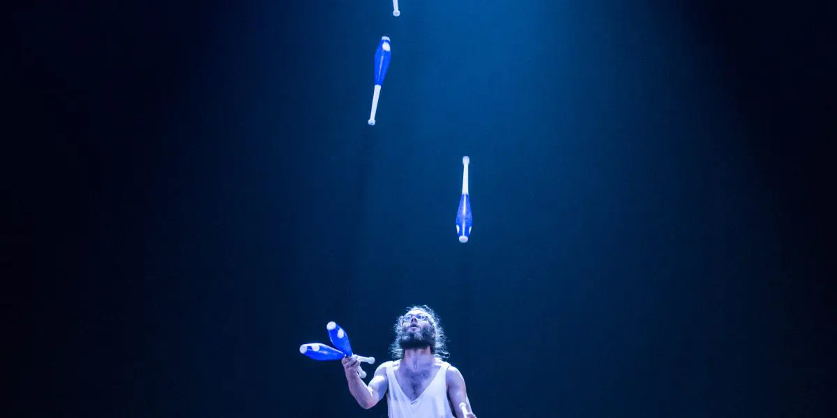 Kellin-Quinn Hentoff-Killian, vêtu d'une tenue blanche, jongle avec six massues bleues sous un projecteur lumineux sur une scène sombre, les massues étant suspendues dans les airs.