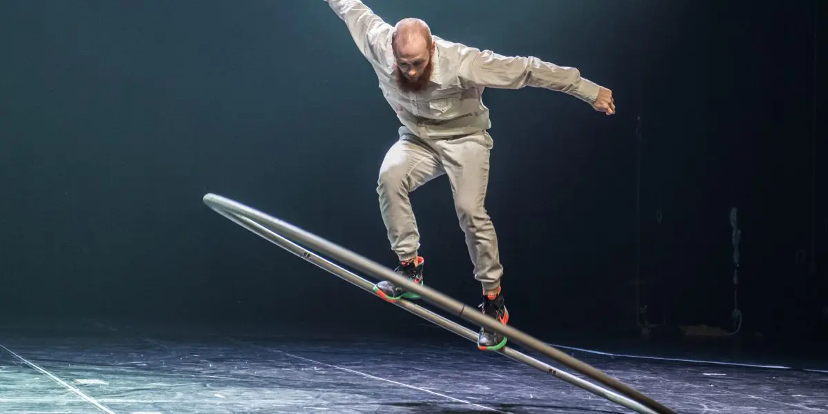 Cooper Smith, vêtu d'une tenue gris clair, se tient habilement en équilibre sur une grande roue Cyr en métal sur une scène faiblement éclairée. Un bras est tendu vers l'extérieur tandis qu'il se penche sur le côté, la roue étant inclinée par rapport au sol.