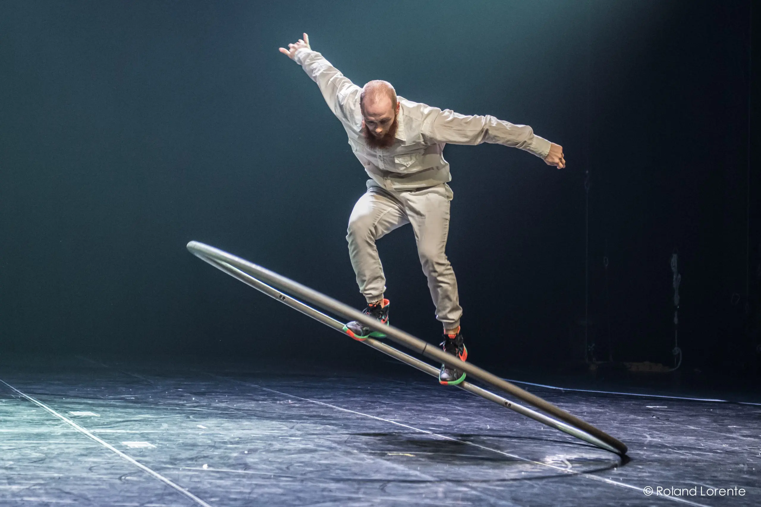 Cooper Smith, vêtu d'une tenue gris clair, se tient habilement en équilibre sur une grande roue Cyr en métal sur une scène faiblement éclairée. Un bras est tendu vers l'extérieur tandis qu'il se penche sur le côté, la roue étant inclinée par rapport au sol.