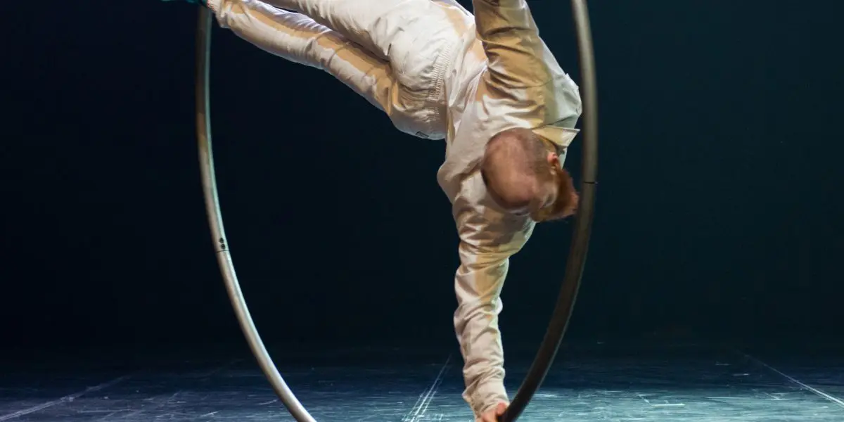 Cooper Smith, vêtu d'une tenue blanche et de chaussures vertes, se tient en équilibre horizontal à l'intérieur d'un grand cerceau métallique (roue Cyr) sur une scène brillante, éclairée sur un fond sombre.