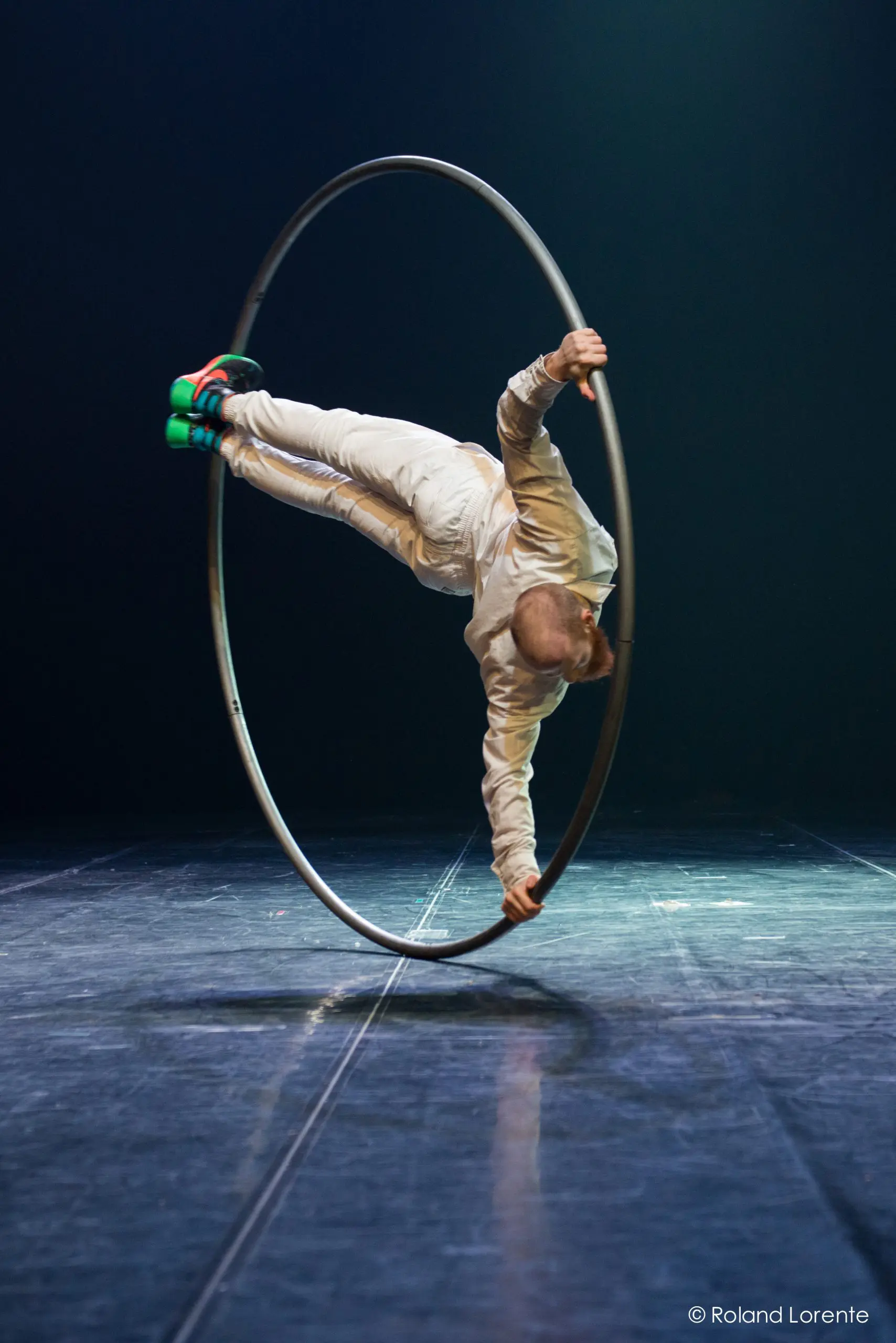 Cooper Smith, vêtu d'une tenue blanche et de chaussures vertes, se tient en équilibre horizontal à l'intérieur d'un grand cerceau métallique (roue Cyr) sur une scène brillante, éclairée sur un fond sombre.