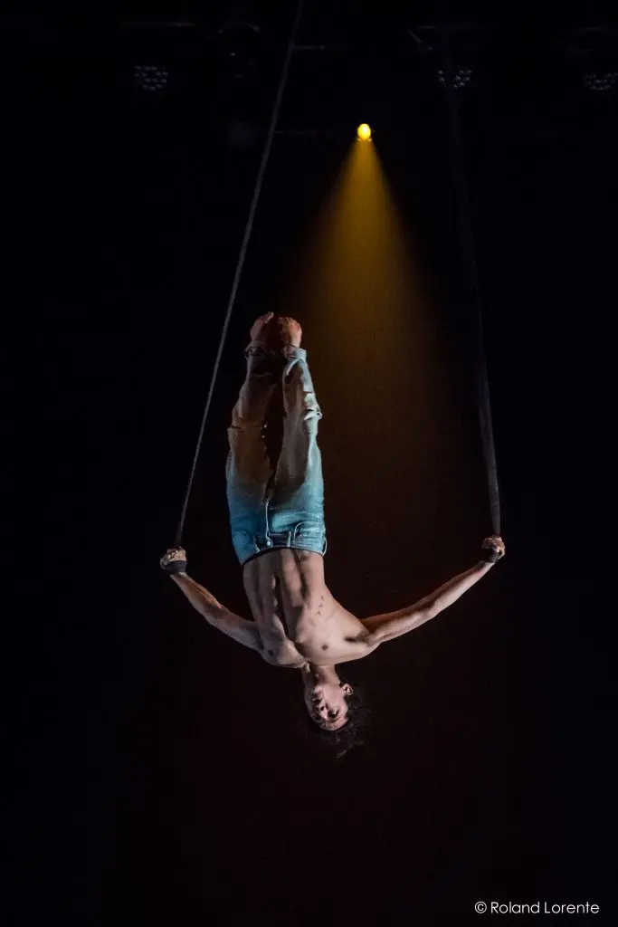 Un artiste aérien, Ricardo Casteneyra, est suspendu à des sangles, les bras gracieusement étendus, sous les projecteurs d'une scène sombre.