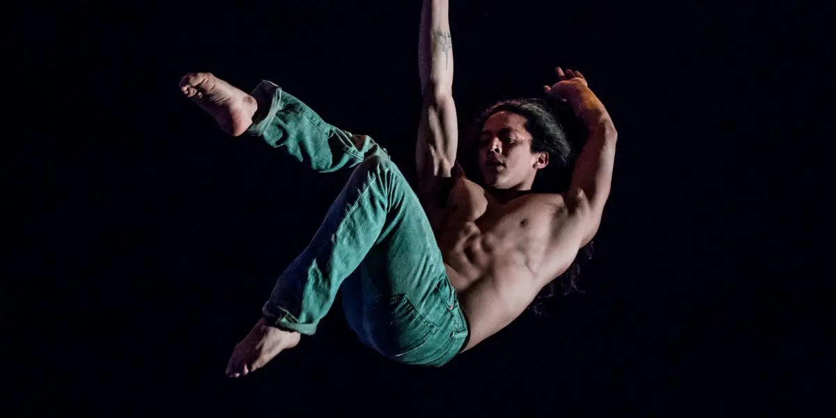 Un artiste aérien torse nu, Ricardo Casteneyra, vêtu d'un pantalon vert, est suspendu à un long tissu, se tenant d'une main tout en étendant ses jambes sur un fond sombre.