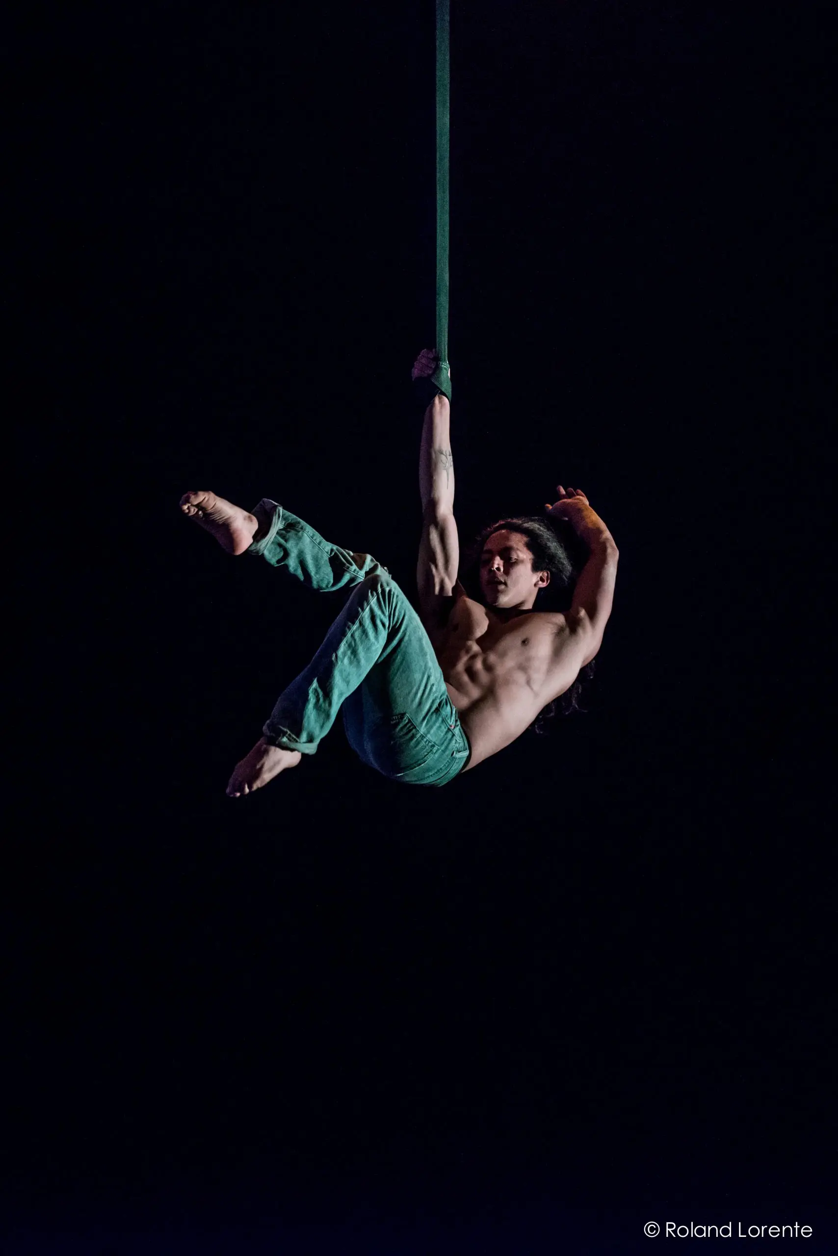 Un artiste aérien torse nu, Ricardo Casteneyra, vêtu d'un pantalon vert, est suspendu à un long tissu, se tenant d'une main tout en étendant ses jambes sur un fond sombre.