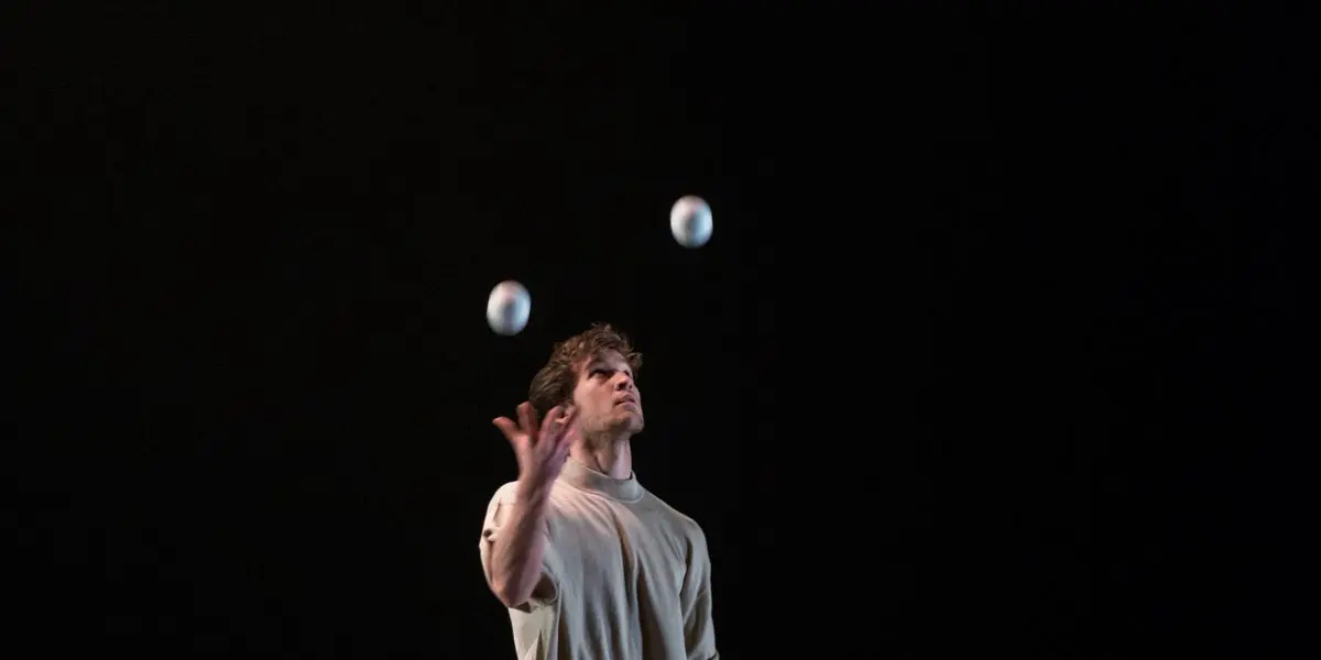 Un homme pieds nus, vêtu d'une chemise grise et d'un pantalon sombre, évoquant Philippe Dupuis, jongle avec sept balles blanches sur une scène sombre, avec deux seaux métalliques en arrière-plan.