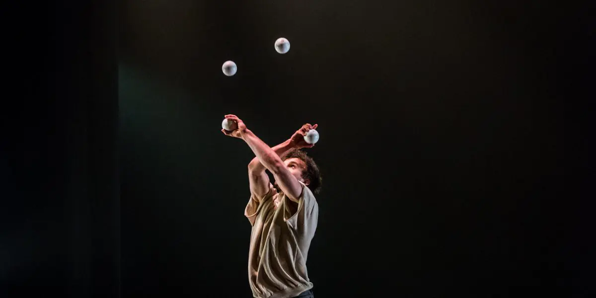 Pieds nus sur scène, Philippe Dupuis jongle avec des balles en croisant les bras. Des seaux en métal sont empilés autour de lui, tandis que des projecteurs projettent des ombres dramatiques sur le fond sombre.