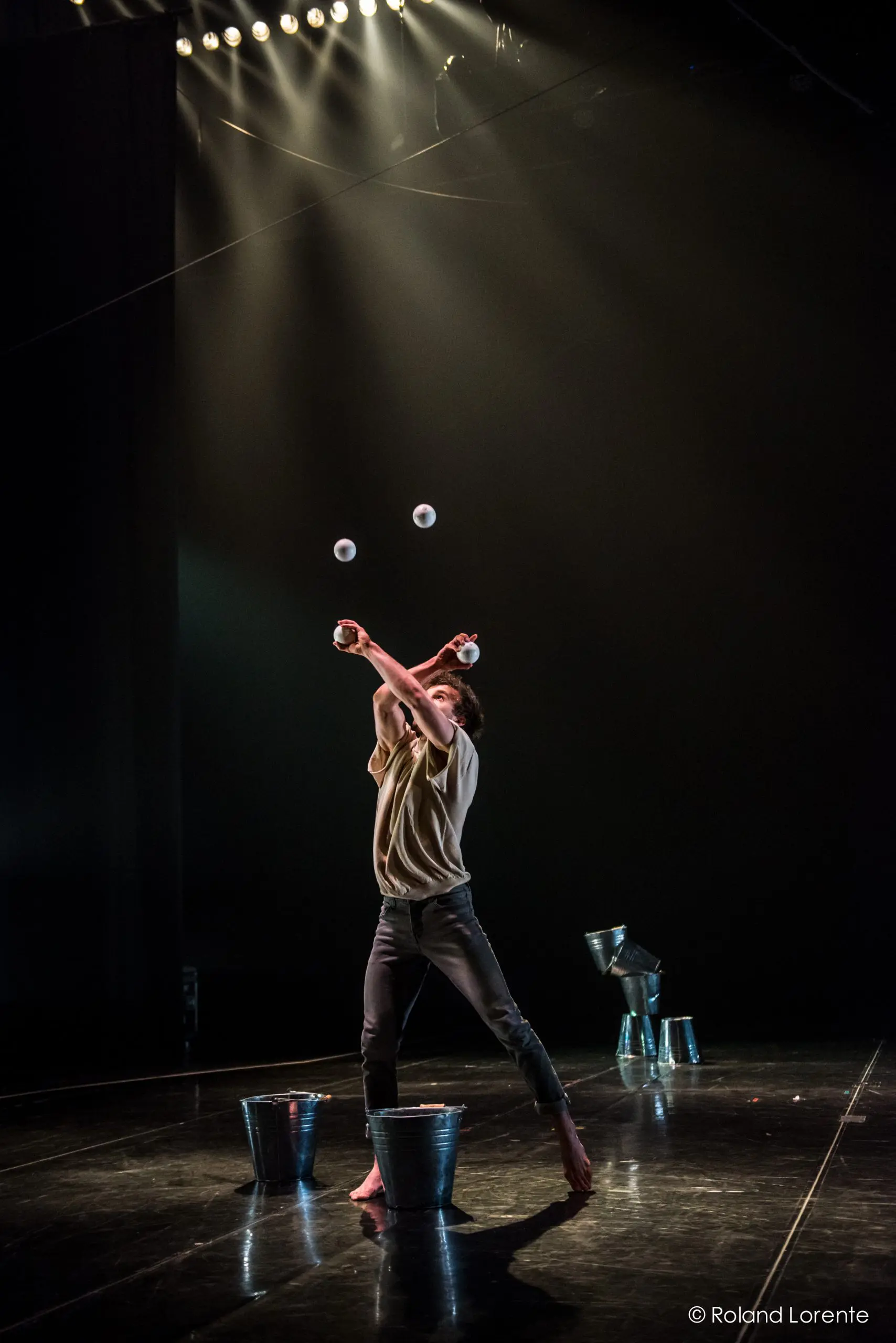 Pieds nus sur scène, Philippe Dupuis jongle avec des balles en croisant les bras. Des seaux en métal sont empilés autour de lui, tandis que des projecteurs projettent des ombres dramatiques sur le fond sombre.