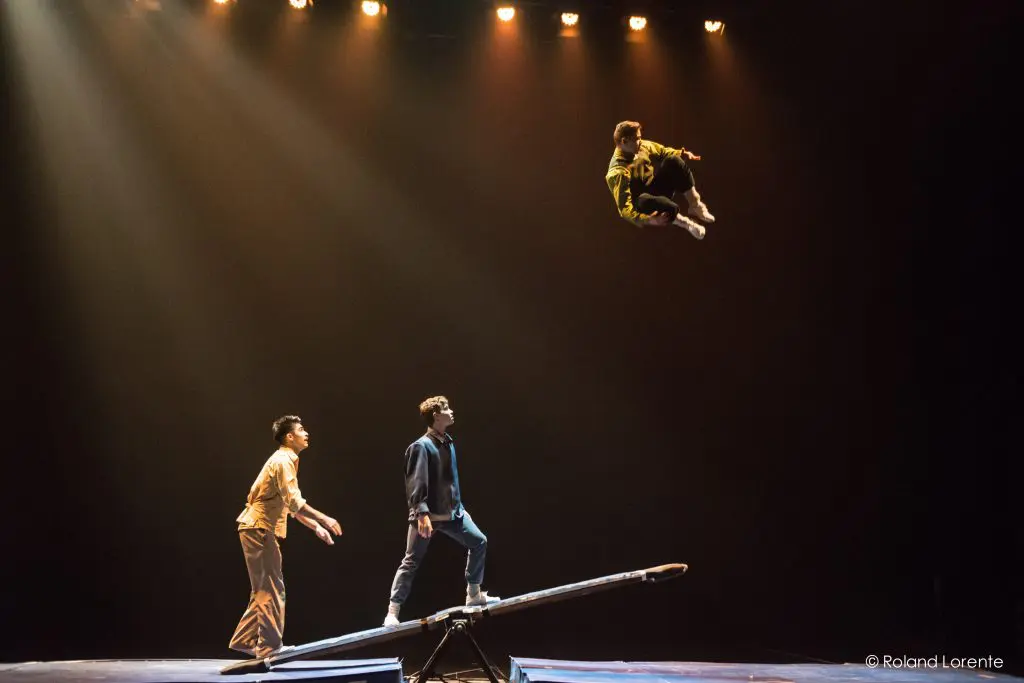 Trois artistes, dont Guillaume Larouche, sont sur scène sous les projecteurs ; l'un d'eux s'élance dans les airs à partir d'une bascule, tandis que les deux autres le regardent attentivement en bas, prêts à l'attraper ou à le soutenir.