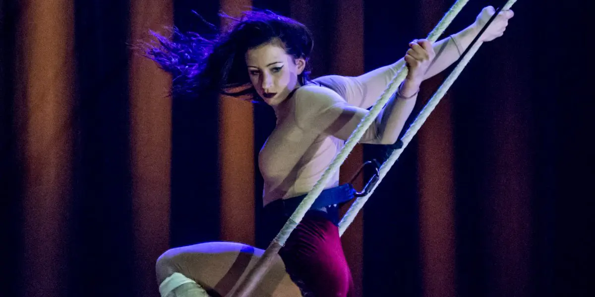 Une voltigeuse aux longs cheveux noirs évolue sur un trapèze, agrippée aux cordes et en équilibre dans les airs sur un fond sombre et éclairé en bleu - une scène qui rappelle l'art captivant de Giulia Scamarcia.