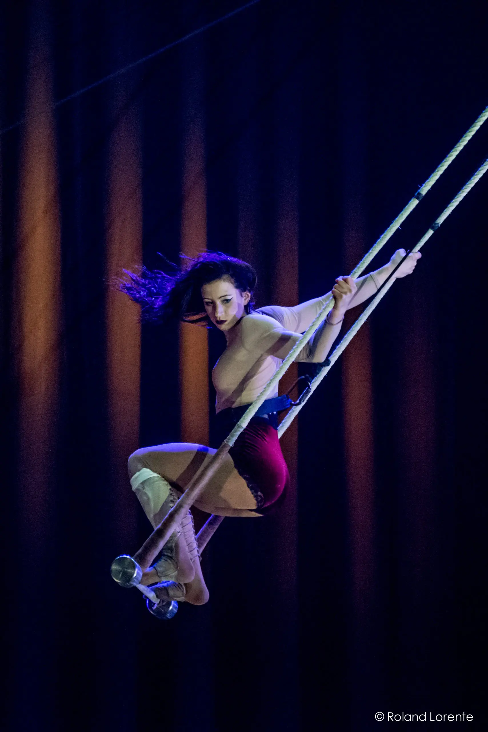 Une voltigeuse aux longs cheveux noirs évolue sur un trapèze, agrippée aux cordes et en équilibre dans les airs sur un fond sombre et éclairé en bleu - une scène qui rappelle l'art captivant de Giulia Scamarcia.