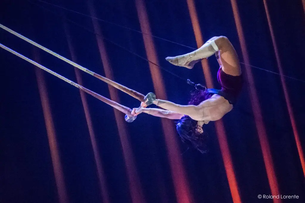 Une trapéziste, ressemblant à Giulia Scamarcia, vêtue d'un short et de chaussettes, est suspendue à l'envers dans les airs en s'accrochant à deux cordes, sur un fond sombre avec des faisceaux lumineux orange en diagonale.