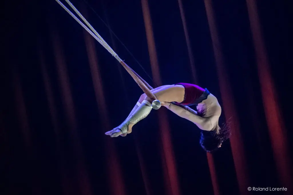 L'acrobate Giulia Scamarcia est suspendue dans les airs, la tête en bas, agrippée à un trapèze avec ses genoux. Elle porte un haut blanc, un short rouge et des bandes blanches aux pieds, et se produit sur un fond sombre avec des faisceaux lumineux orange spectaculaires.