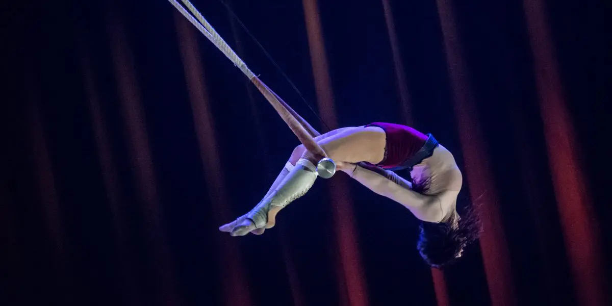 L'acrobate Giulia Scamarcia est suspendue dans les airs, la tête en bas, agrippée à un trapèze avec ses genoux. Elle porte un haut blanc, un short rouge et des bandes blanches aux pieds, et se produit sur un fond sombre avec des faisceaux lumineux orange spectaculaires.