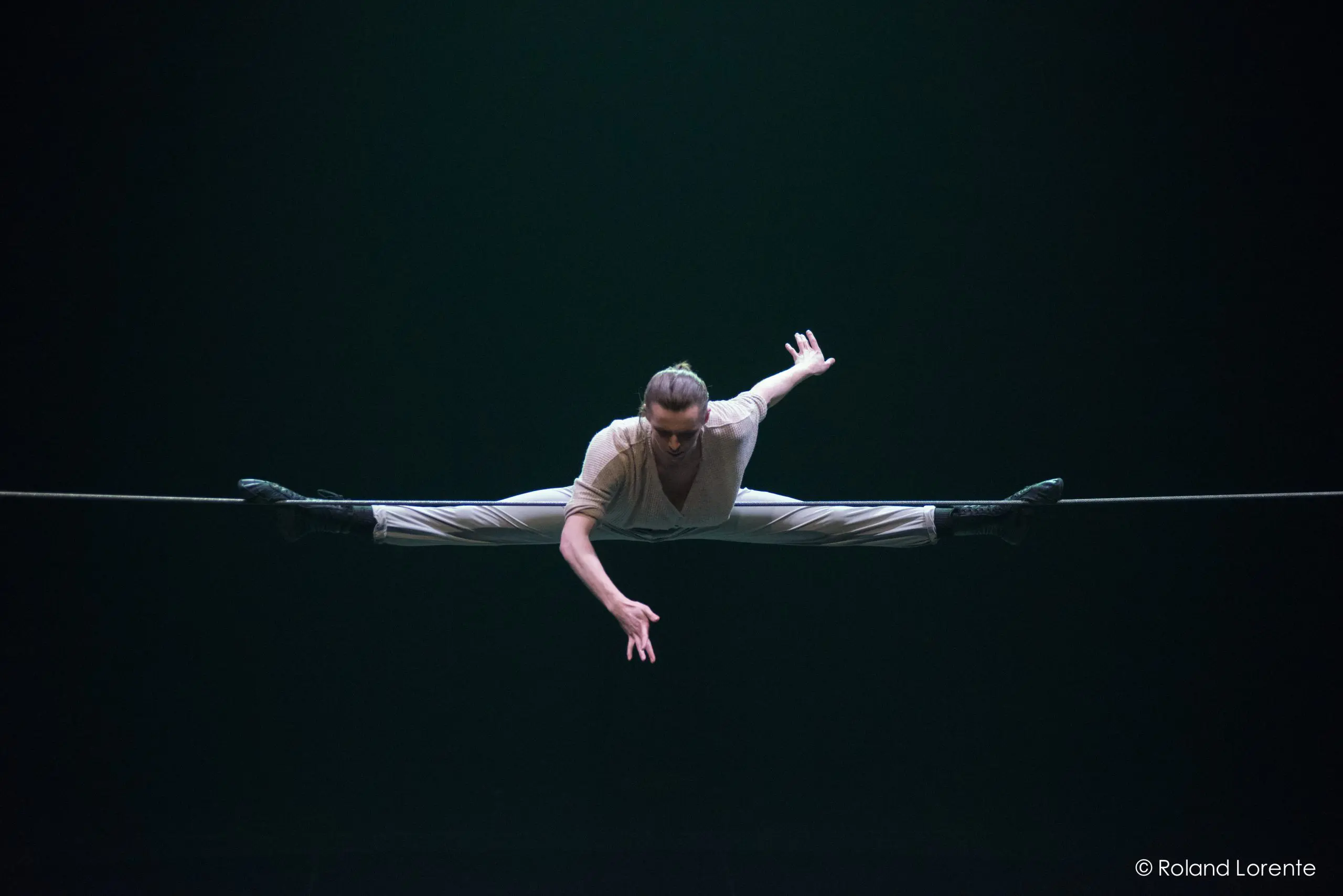 Un artiste se tient en équilibre sur une corde raide sous les feux de la rampe, faisant le grand écart avec les bras gracieusement étendus et les jambes tendues à l'horizontale sur la corde.