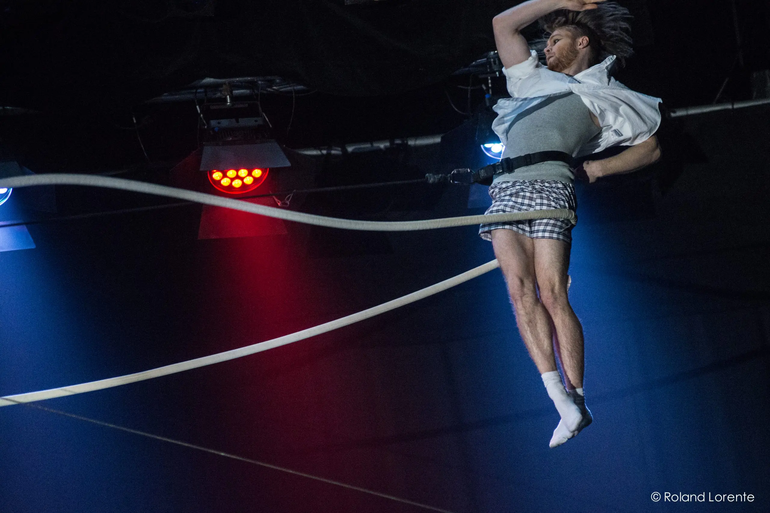 Nathan Jones, un acrobate masculin vêtu d'un short à carreaux et d'une chemise blanche, exécute une acrobatie en plein vol sur une slackline sous un éclairage scénique spectaculaire, sa chemise soulevée par le mouvement et un bras levé au-dessus de sa tête.
