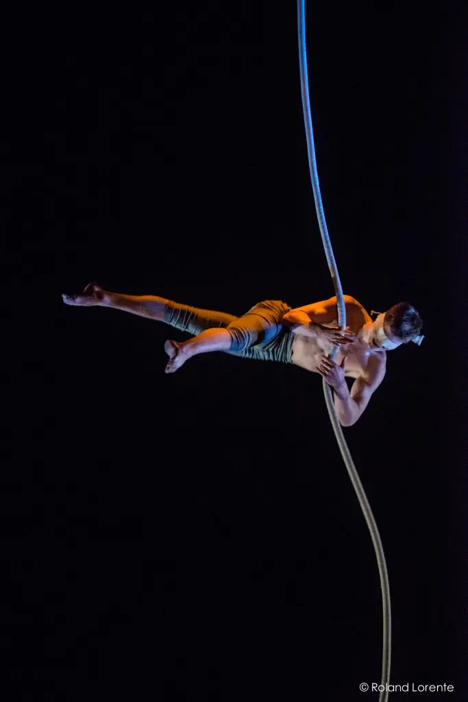 Maxime Piché-Luneau, un artiste aérien, est suspendu horizontalement dans les airs, torse nu et vêtu d'un pantalon, agrippé à une corde verticale sur un fond sombre.