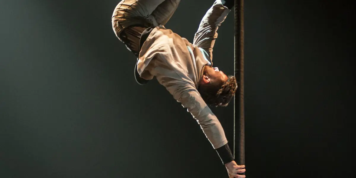 Raphaël Filiatreault se tient en équilibre à l'envers sur un poteau vertical, qu'il saisit d'une main et des deux jambes, tandis que son corps se courbe et qu'un bras s'étend vers le bas, le tout sous un éclairage scénique dramatique.
