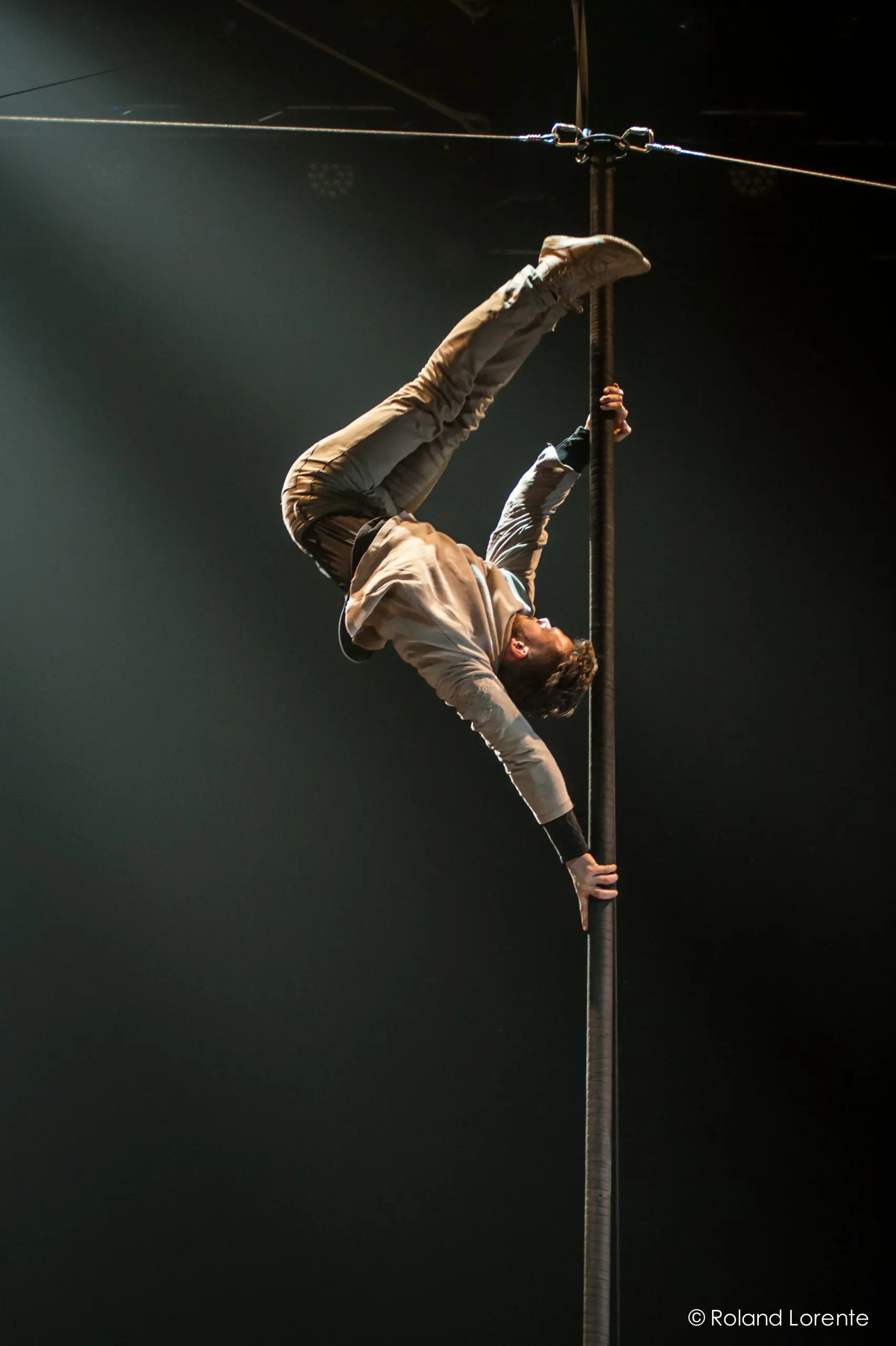Raphaël Filiatreault se tient en équilibre à l'envers sur un poteau vertical, qu'il saisit d'une main et des deux jambes, tandis que son corps se courbe et qu'un bras s'étend vers le bas, le tout sous un éclairage scénique dramatique.
