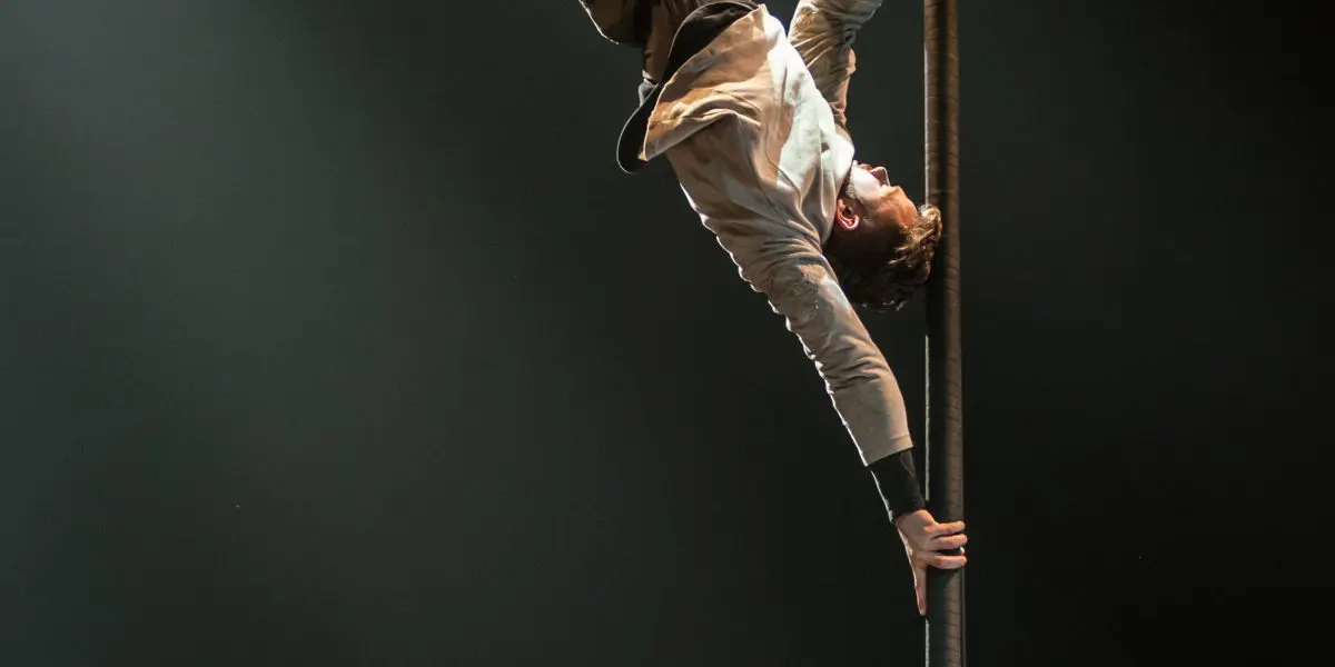 Un artiste de cirque, comme Raphaël Filiatreault, se tient en équilibre horizontal avec une main sur un poteau vertical sous un projecteur, les jambes étendues et le corps parallèle au sol, dans une salle obscure.