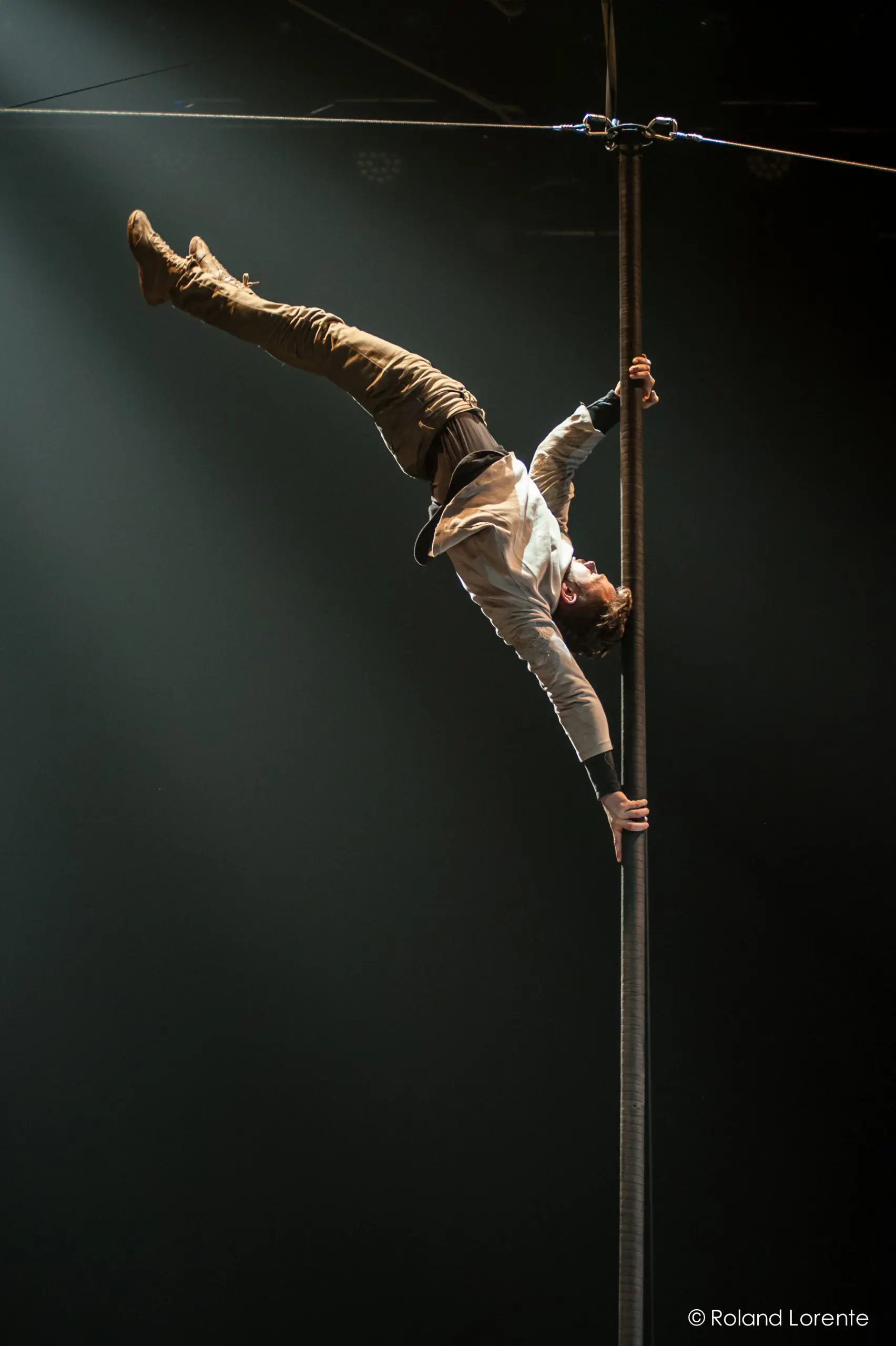 Un artiste de cirque, comme Raphaël Filiatreault, se tient en équilibre horizontal avec une main sur un poteau vertical sous un projecteur, les jambes étendues et le corps parallèle au sol, dans une salle obscure.