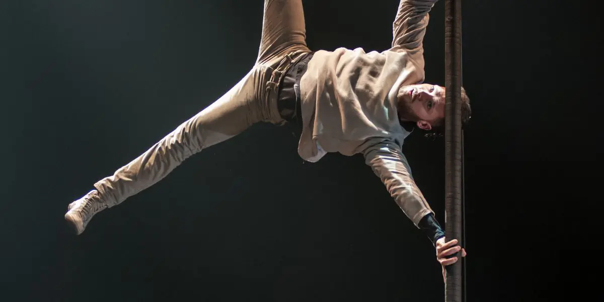 Un homme en tenue décontractée exécute un mouvement acrobatique impressionnant, suspendu à l'horizontale, une main agrippée à un poteau vertical, les jambes écartées, éclairé par les projecteurs sur une scène sombre, faisant preuve d'une agilité qui rappelle celle de Raphaël Filiatreault.