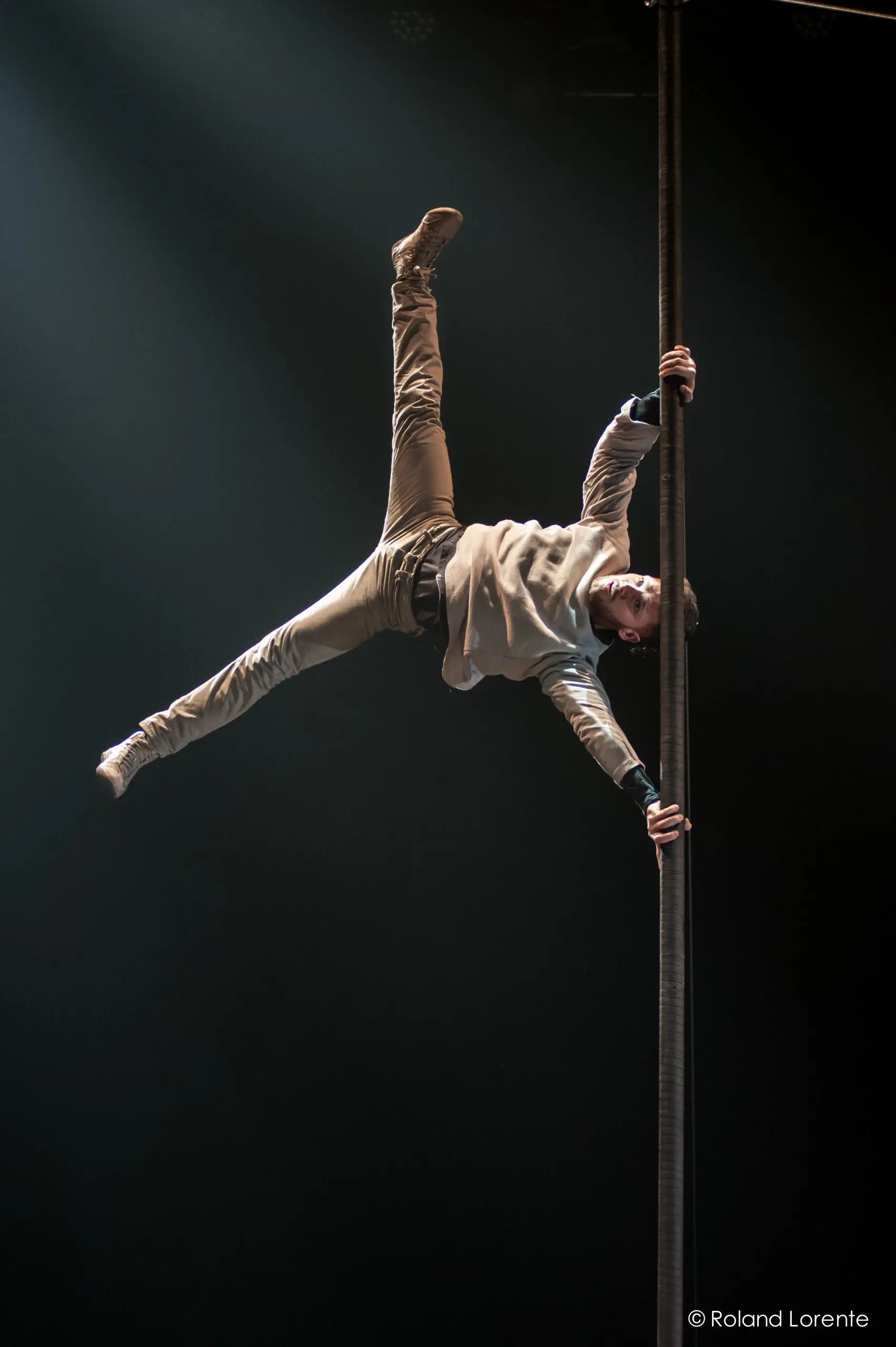 Un homme en tenue décontractée exécute un mouvement acrobatique impressionnant, suspendu à l'horizontale, une main agrippée à un poteau vertical, les jambes écartées, éclairé par les projecteurs sur une scène sombre, faisant preuve d'une agilité qui rappelle celle de Raphaël Filiatreault.