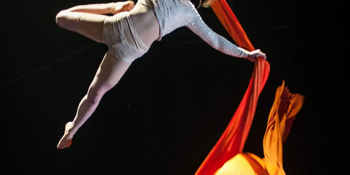 Une voltigeuse, Sabine Van Rensburg, évolue dans les airs, tenant des soies orange vif d'une main et d'une jambe tendue sur un fond sombre. Sa tenue blanche capte la lumière tandis que le tissu s'écoule de manière spectaculaire sous elle.