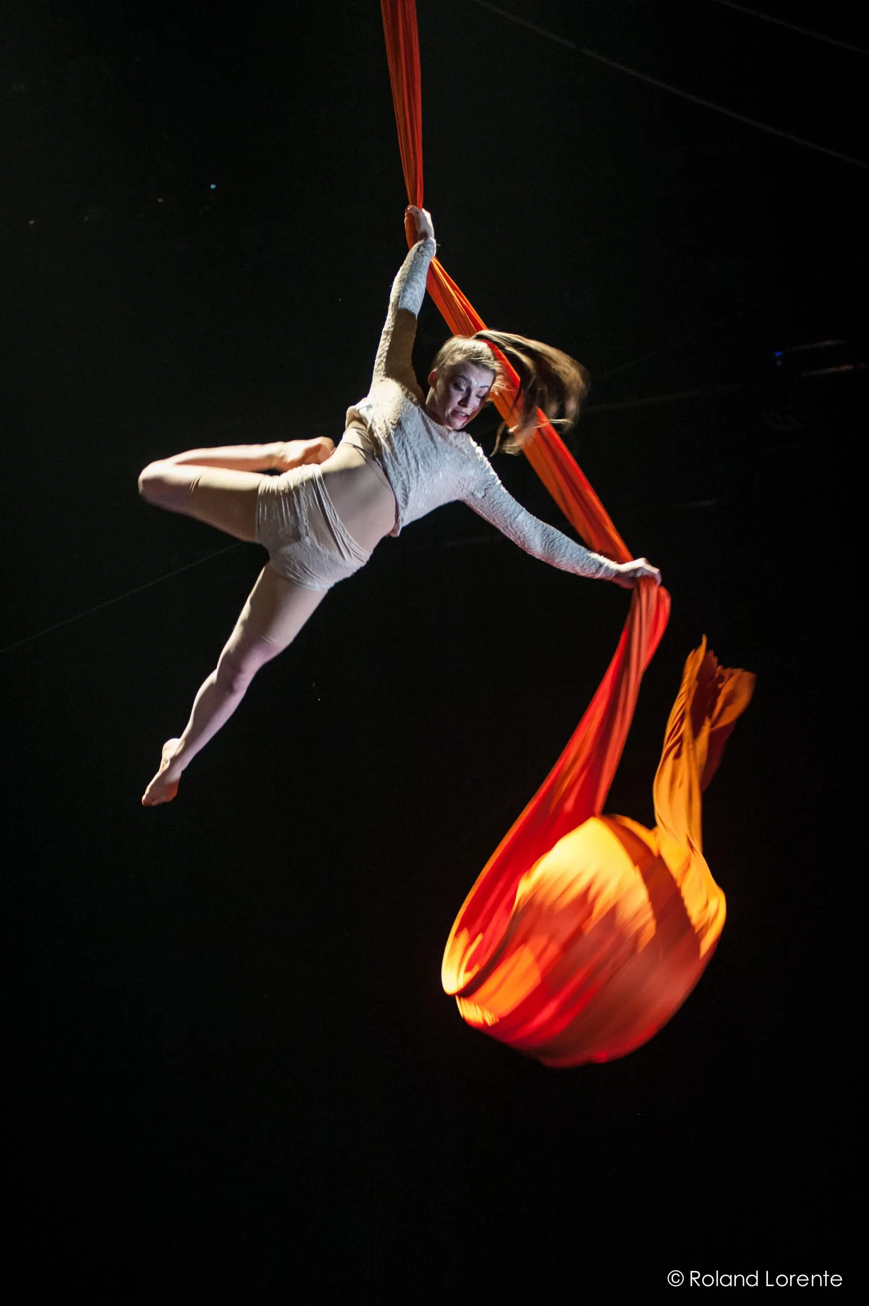 Une voltigeuse, Sabine Van Rensburg, évolue dans les airs, tenant des soies orange vif d'une main et d'une jambe tendue sur un fond sombre. Sa tenue blanche capte la lumière tandis que le tissu s'écoule de manière spectaculaire sous elle.