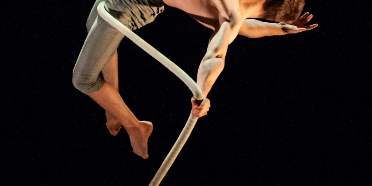 Maxime Piché-Luneau, un artiste aérien torse nu, en pantalon léger, se penche gracieusement vers l'arrière tout en étant suspendu à une corde sur un fond noir. Un bras est tendu tandis que l'autre tient la corde.