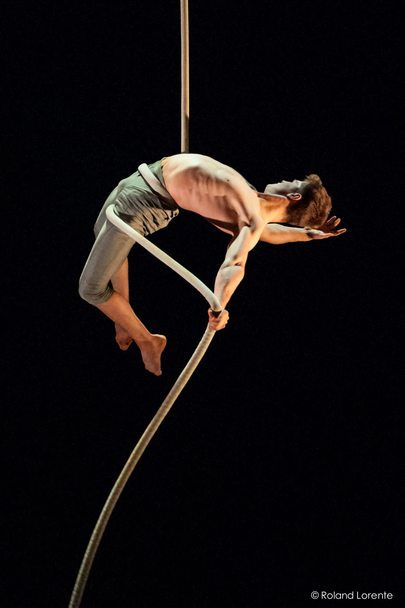 Maxime Piché-Luneau, un artiste aérien torse nu, en pantalon léger, se penche gracieusement vers l'arrière tout en étant suspendu à une corde sur un fond noir. Un bras est tendu tandis que l'autre tient la corde.