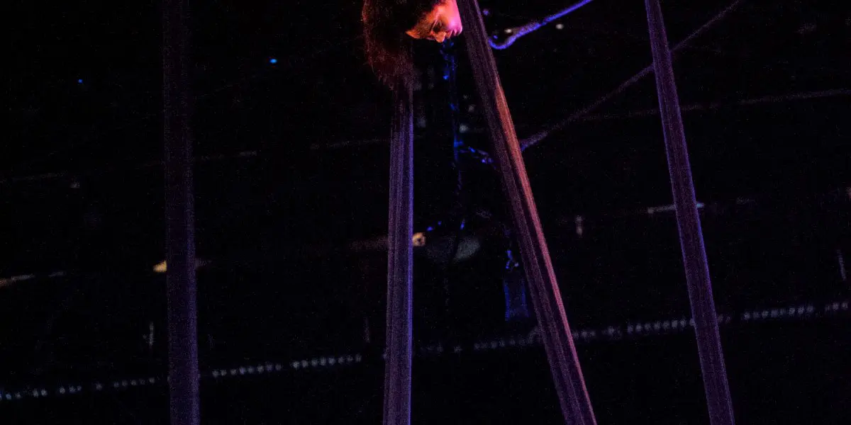 L'artiste aérienne Vanessa Aviles exécute une pose acrobatique en hauteur, suspendue à l'envers par de longues soies sombres sous l'éclairage de la scène.