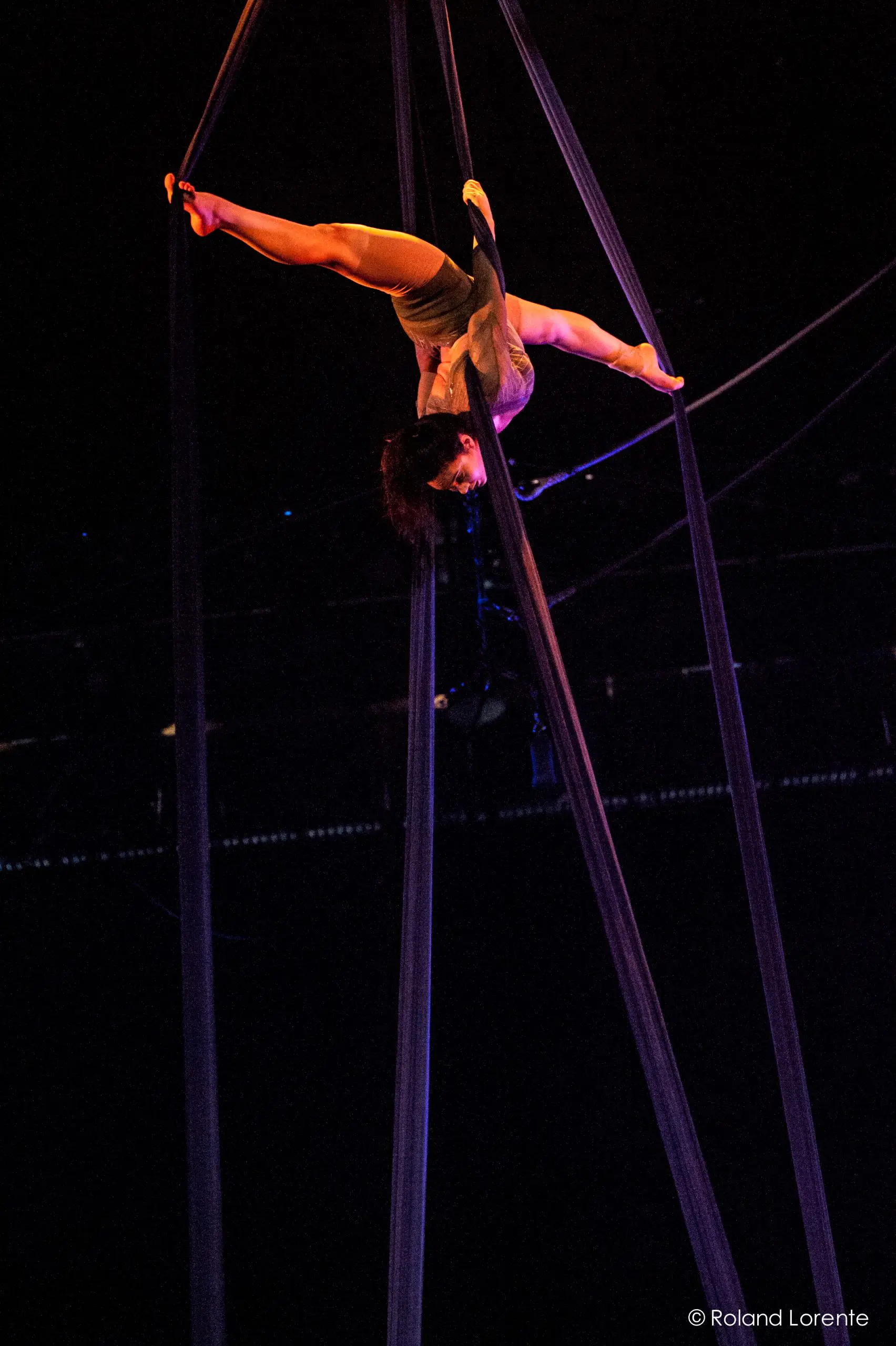 L'artiste aérienne Vanessa Aviles exécute une pose acrobatique en hauteur, suspendue à l'envers par de longues soies sombres sous l'éclairage de la scène.