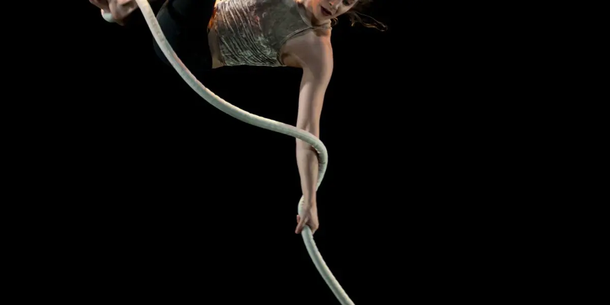 Une voltigeuse, Una Bennett, exécute une routine acrobatique sur une corde blanche, suspendue en l'air sur un fond noir, le corps allongé et les cheveux flottants.