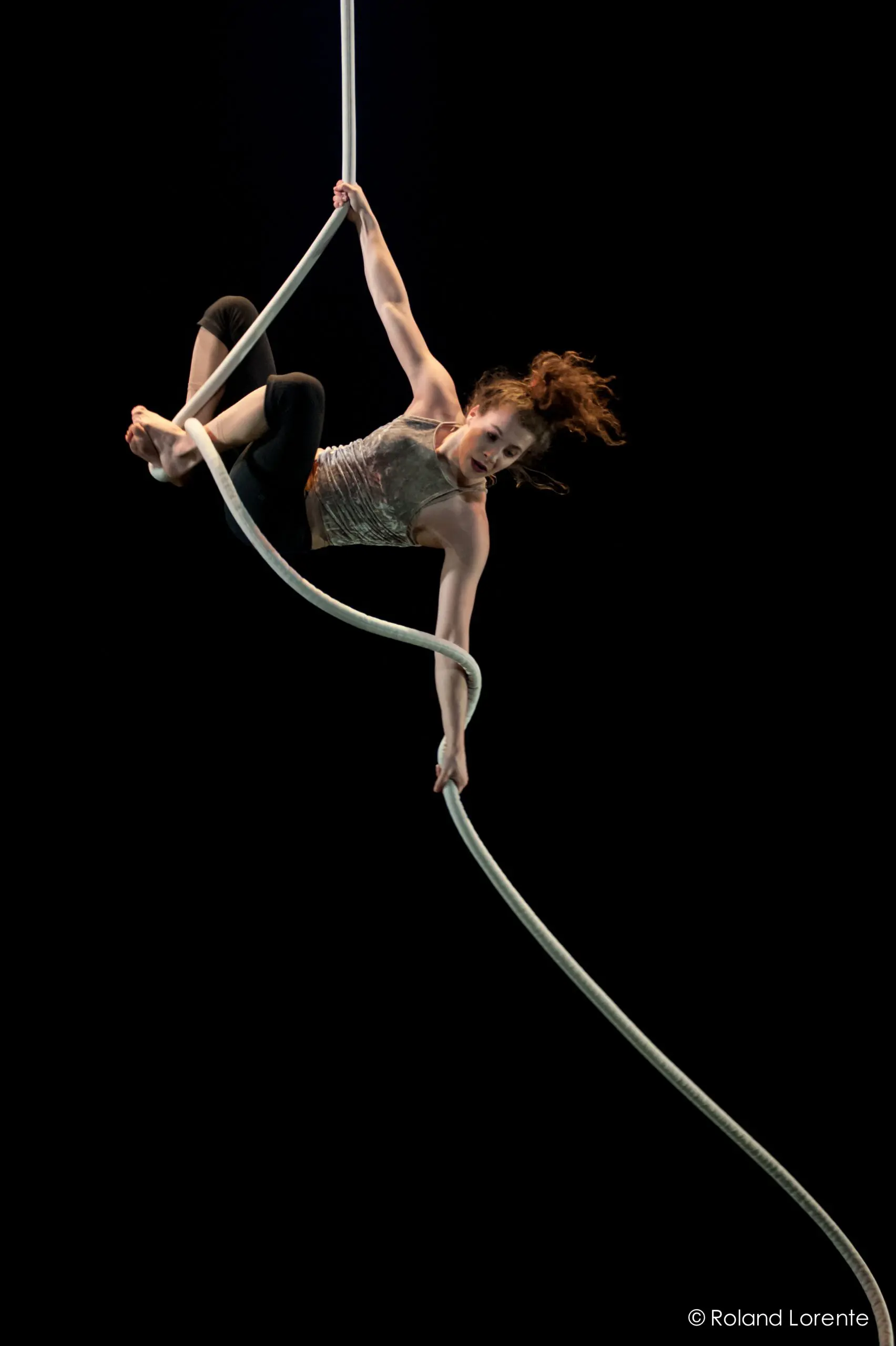 Une voltigeuse, Una Bennett, exécute une routine acrobatique sur une corde blanche, suspendue en l'air sur un fond noir, le corps allongé et les cheveux flottants.
