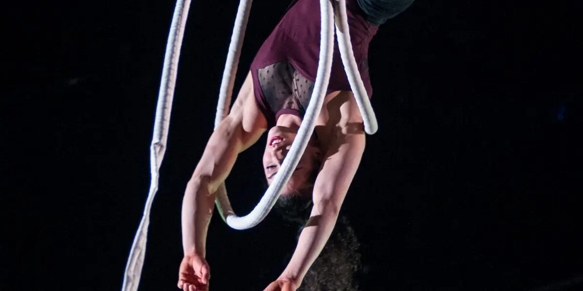 Une artiste de cirque, Rosita Hendry, exécute un numéro aérien, suspendue la tête en bas à une épaisse corde blanche, les jambes et les bras gracieusement étendus, sur un fond sombre.