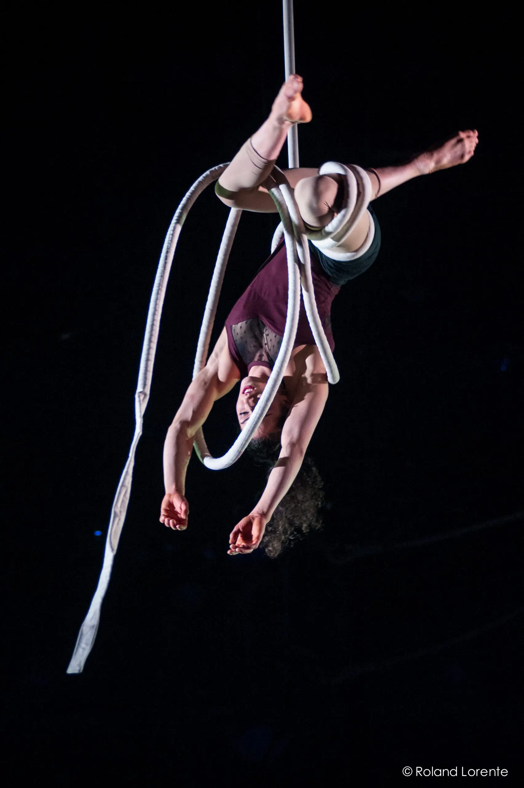 Une artiste de cirque, Rosita Hendry, exécute un numéro aérien, suspendue la tête en bas à une épaisse corde blanche, les jambes et les bras gracieusement étendus, sur un fond sombre.